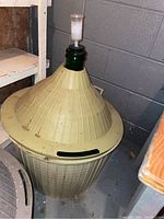 Green glass demijohn inside beige plastic protective outer case with removable lid and fitted airlock stopper on top, photographed in basement setting.