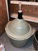 Green glass demijohn inside gray plastic protective basket with handles, topped with cork and airlock, placed in basement storage.