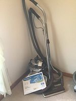 Filter Queen vacuum with hose and metal wand standing in a corner next to two packages of cone filters on the floor; power head floor tool attached