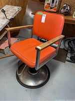 Front angle of orange vinyl barber chair showing padded seat and backrest with wooden armrests and rusty metal base.