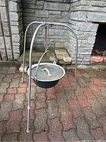 Full view of the Hungarian Bogrács cooking pot suspended from the stainless steel tripod stand on outdoor paved surface with stone wall in background.