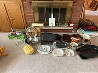 Photo showing a range of kitchenware arranged on carpet in front of fireplace, including Dutch oven, roasting pan, baking dishes, mixing bowls, cutting boards, and vintage electric utensil.