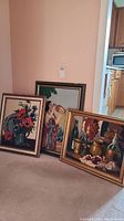 Three framed needlepoint art pieces leaning against a wall on carpet, showing a floral piece, woman drinking, and wine still life.
