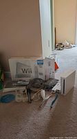 Wide shot showing Nintendo Wii console, original box, two Wii remotes with wrist straps and silicone covers, Nunchuck joystick, sensor bar, power adapter, manuals and game disc scattered on carpet.