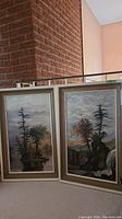 Pair of framed oil paintings showing trees and natural landscapes, placed leaning against a brick wall.