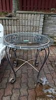 Full view of round patio table showing clear glass top and decorative metal base with curved legs and cross supports, sitting on outdoor patio surface.