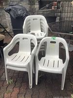 Photo showing three white resin lawn chairs placed on a paved surface with a brick wall backdrop.