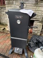 Front view of assembled smoker with instruction manual on top, propane tank and cover bag beside it