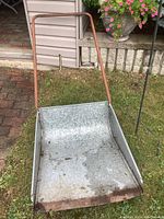 Full view of aluminum snow scoop with metal handle placed on grass and stone patio, showing overall condition and size.