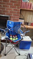 Blue outbound folding canvas chair holding a Head Ti.Evolution tennis racket, three cans of Penn tennis balls, next to a blue and white Coleman cooler and brick fireplace.