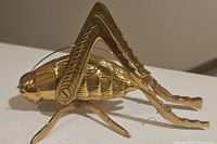 Side view of shiny brass grasshopper figurine highlighting the detailed legs and textured body.