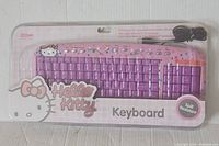 Front view of purple Hello Kitty themed USB keyboard in clear plastic packaging showing full QWERTY key layout and multimedia keys.