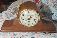 Full front view of the wooden mantle clock on a floral cloth. Shows the clock's curved wooden case and white dial with black numerals and hands.