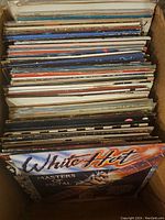 Box filled with numerous vintage vinyl records in sleeves showing varied titles and colors.