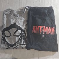 Both folded t-shirts laid side by side on a white surface, showing the gray Funko Pop! Spider-Man shirt and the black Ant-Man shirt.