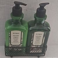 Two green pump bottles labeled Holiday Forest, one hand lotion and one hand wash, placed in a black mesh holder.