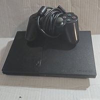 Black Sony PlayStation 2 Slim console with wired controller resting on top, showing the front and top side with buttons and logos visible.