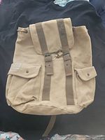 Front view of the tan canvas military-style backpack with flap top and buckle straps, showing two front button flap pockets.