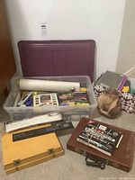 Plastic storage containers filled with assorted art supplies including sketch pads, markers, pencils, and tools with a carved figure beside.