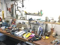 Wide shot of workbench with assortment of hand tools and hardware on surface and shelves showing items including hammers, screwdrivers, utility knives, small parts containers, and packaged accessories.