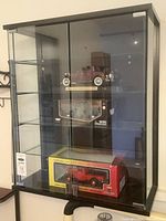 Front view of black glass display case with glass shelves showing the three diecast model cars inside.