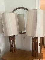 Two wood based lamps with beige cylindrical shades placed on a wooden surface, with a round wood framed mirror hanging on the wall behind them.