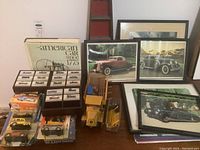 Multiple small diecast cars in a wooden display case, vintage style truck model with wooden crates and barriers, framed vintage car prints, and an automotive book titled 'The American Car Since 1775'.