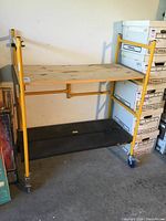 Full view of the yellow metal mobile scaffolding showing the wooden top platform and black base shelf on four caster wheels