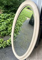 Oval wall mirror leaning on the ground outdoors showing cream chipped paint frame and clear reflective glass surface.
