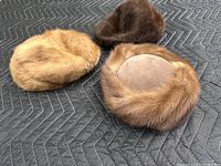 Three fur pillbox hats laid out on a dark quilted surface, showing variations in color and texture.
