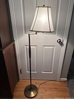 Full view of a mid-century modern swing arm floor lamp with white fabric trapezoidal shade, brass and wood components, standing against a white door.