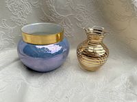 Two vases displayed on fabric background: one iridescent purple with gold band, one clear glass with gold stripes and flared opening.