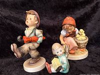 Photo showing all three Goebel Hummel figurines together: Globe Trotter carrying basket and umbrella, Chick Girl seated with chicks, and Angel with accordion.