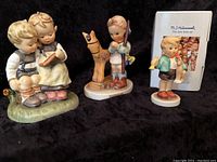 Three Hummel figurines displayed with original box on a black cloth background: two children reading on a bench, a boy with a horse toy, and a boy with a candle.