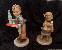 Photo of both Goebel Hummel figurines side by side: Waiter boy holding tray with wine bottle and glass, and girl doll bathing smaller doll in tub.