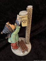 Side view of the Goebel Hummel figurine showing a child in winter clothing placing a letter into a mailbox mounted on a wooden post, with a sled on the base.