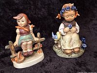 Photo of two Hummel figurines, one sitting on a fence with a bird on base, the other holding a blue flower with a bird next to her.