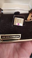 Front view of gold tie clip with engraved ITT logo and red gemstone on black velvet background inside case.