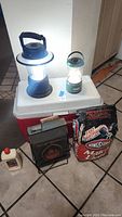 Photo showing Coleman lantern illuminated on top of Coleman ice chest with Utilitech lantern, Son of Hitachi grill and Kingsford charcoal bag visible on floor