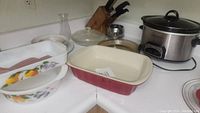 Photo showing Fire King baking dishes with fruit design, a Good Cook ceramic baking dish, and a Crock-Pot slow cooker on the kitchen counter.