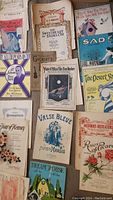 Photo showing an assortment of full size vintage sheet music covers featuring various songs such as The Rose of No Man's Land, The Desert Song, Sad, Sweetheart of Sigma Chi, and others, all aged and worn.