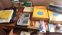 Photo showing a variety of office supplies on a wooden surface including unopened boxes of Staples hanging file folders, two calculators, boxes of envelopes, pens, and other small supplies.