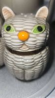 Close-up of ceramic cat-shaped salt or pepper shaker showing textured stripes, green eyes, and an orange nose with brown collar.