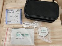 Photo of the Dr. Ho's TENS device, flexible electrode pads sealed in plastic, white connector cord, and black nylon travel bag on wooden surface.