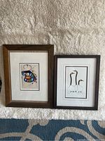 Photo showing both framed prints side by side on carpeted floor with textured white wall background.