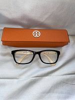 Tory Burch black framed glasses placed below an orange hard case with Tory Burch logo, on white fabric background.