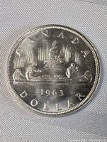 Front side of 1963 Canadian silver dollar coin showing Indigenous person and voyageur paddling canoe with 'CANADA DOLLAR 1963' inscription.