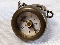 Close-up photo of antique Marbles military compass showing compass needle pointing SE, brass casing and knurled edge with attached metal ring.