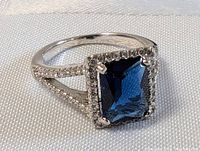 Close-up of sterling silver ring with square deep blue faceted gemstone surrounded by clear stones on split shank band, set on a white fabric background.