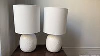 Photo of two white ceramic table lamps with textured bases and white fabric shades on a wooden surface.